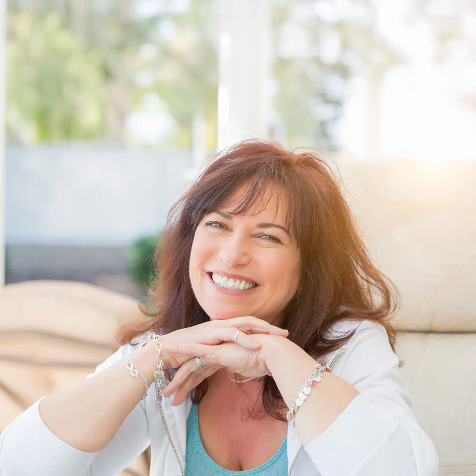 Smiling woman with red hair sitting comfortably outdoors on a cushioned chair in soft sunlight.