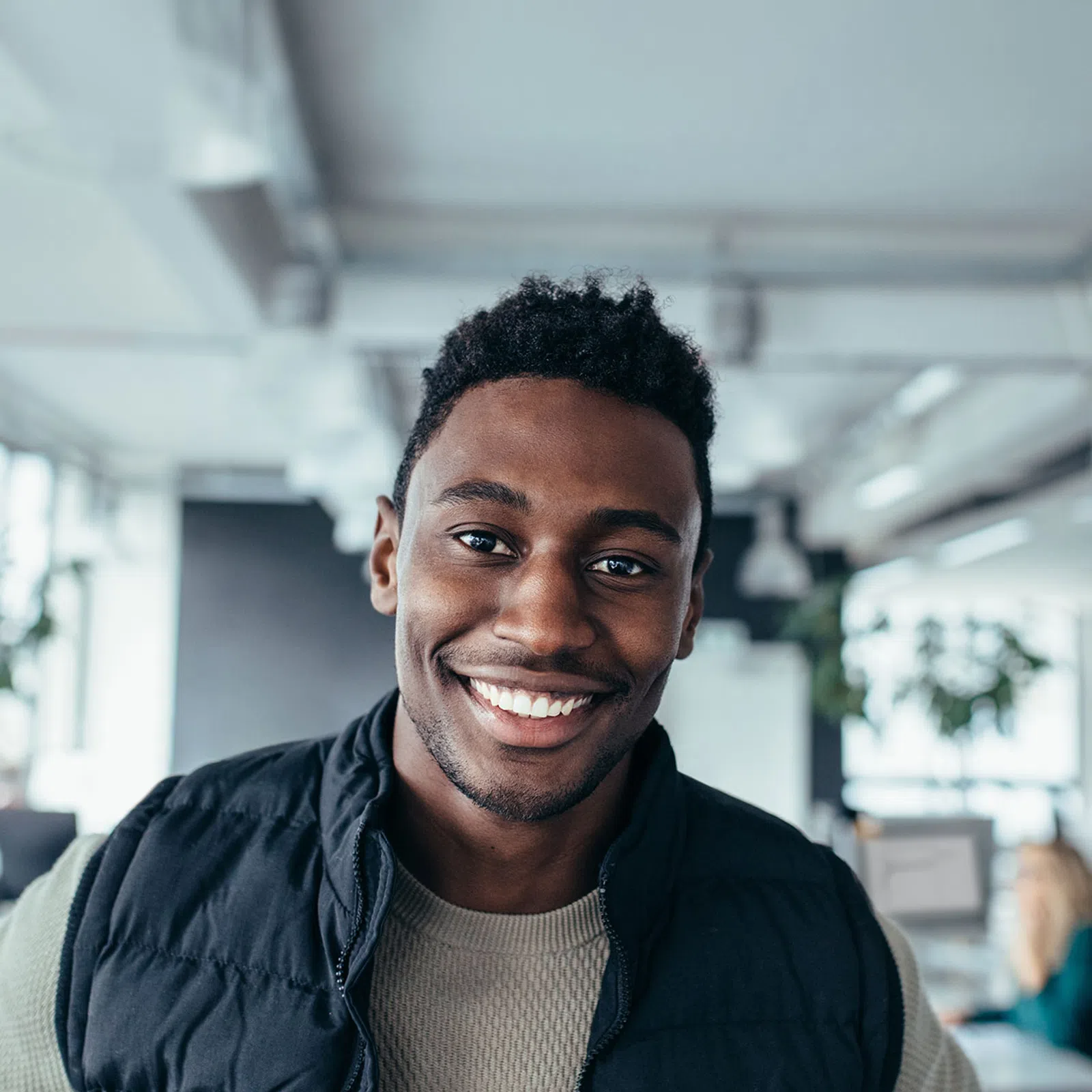 Smiling young man in casual attire standing in a modern office with colleagues working in the background