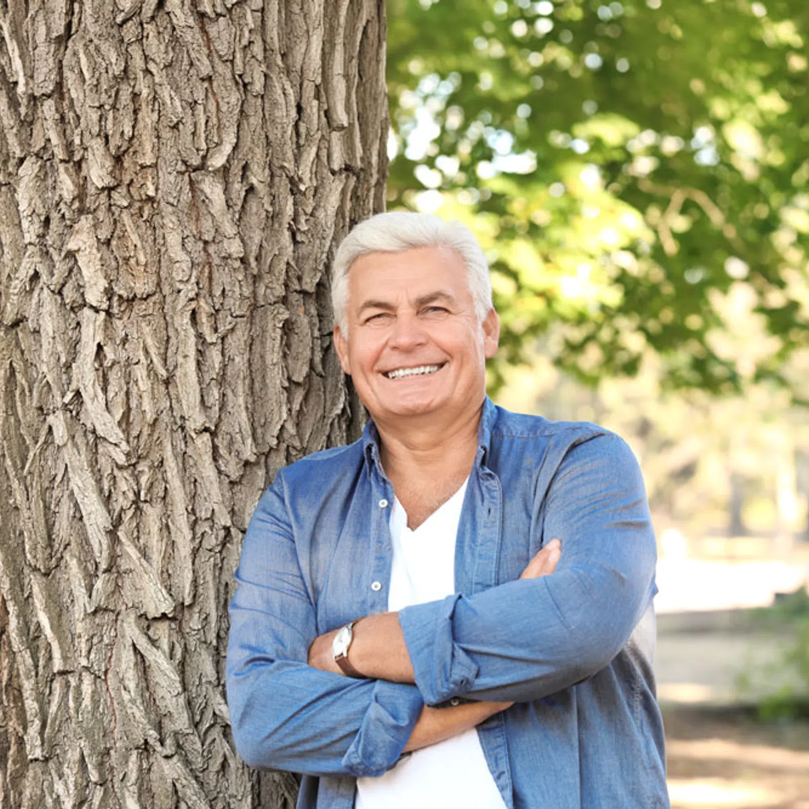Smiling elderly man with white hair leans against a large tree trunk in a sunlit park with green foliage.