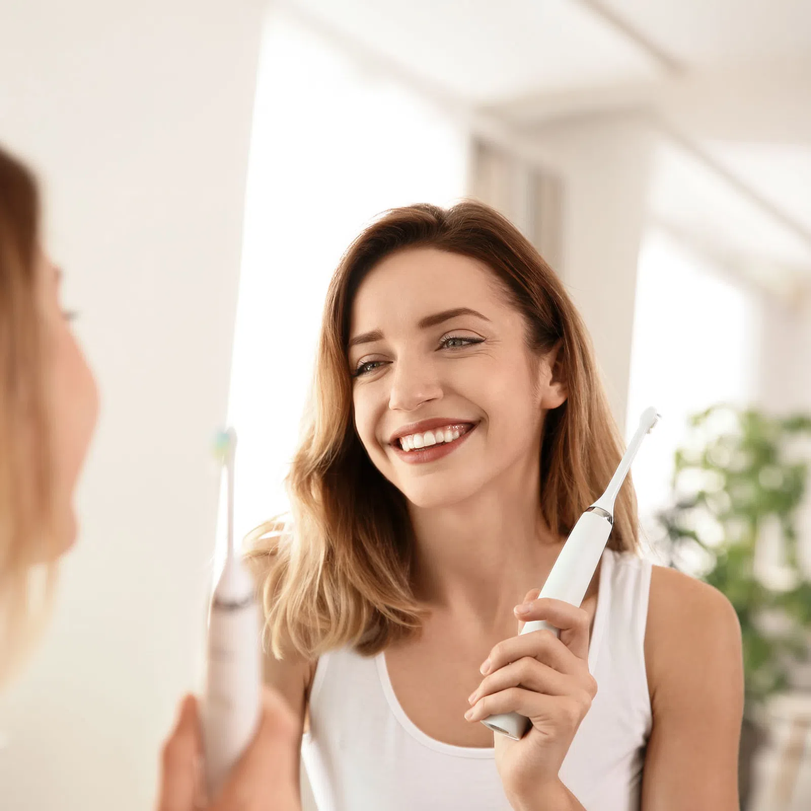 Smiling woman holding electric toothbrush in bathroom mirror, promoting dental hygiene and oral care routine.