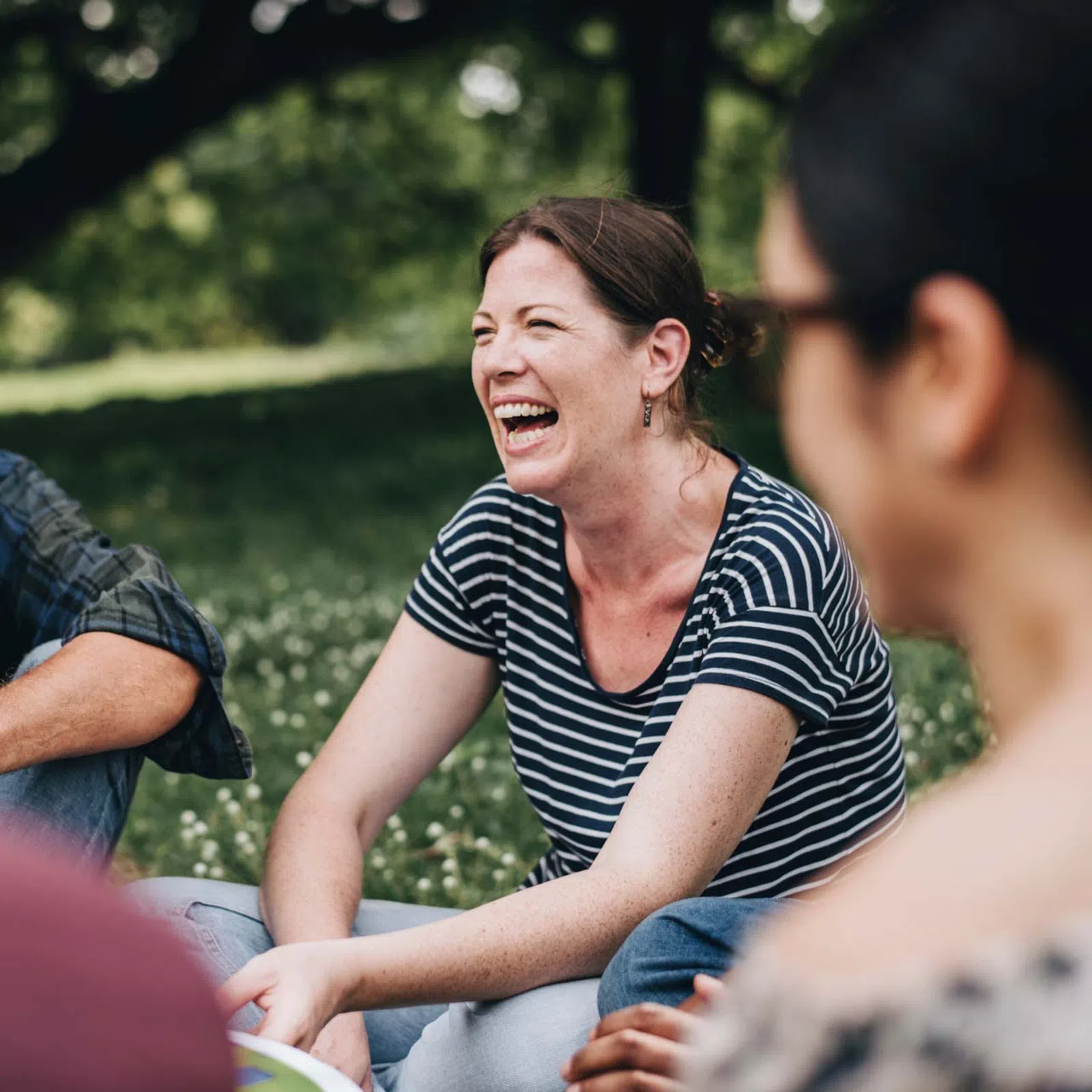 Group of people sitting outdoors on grass, smiling and enjoying a lively conversation in a park setting.