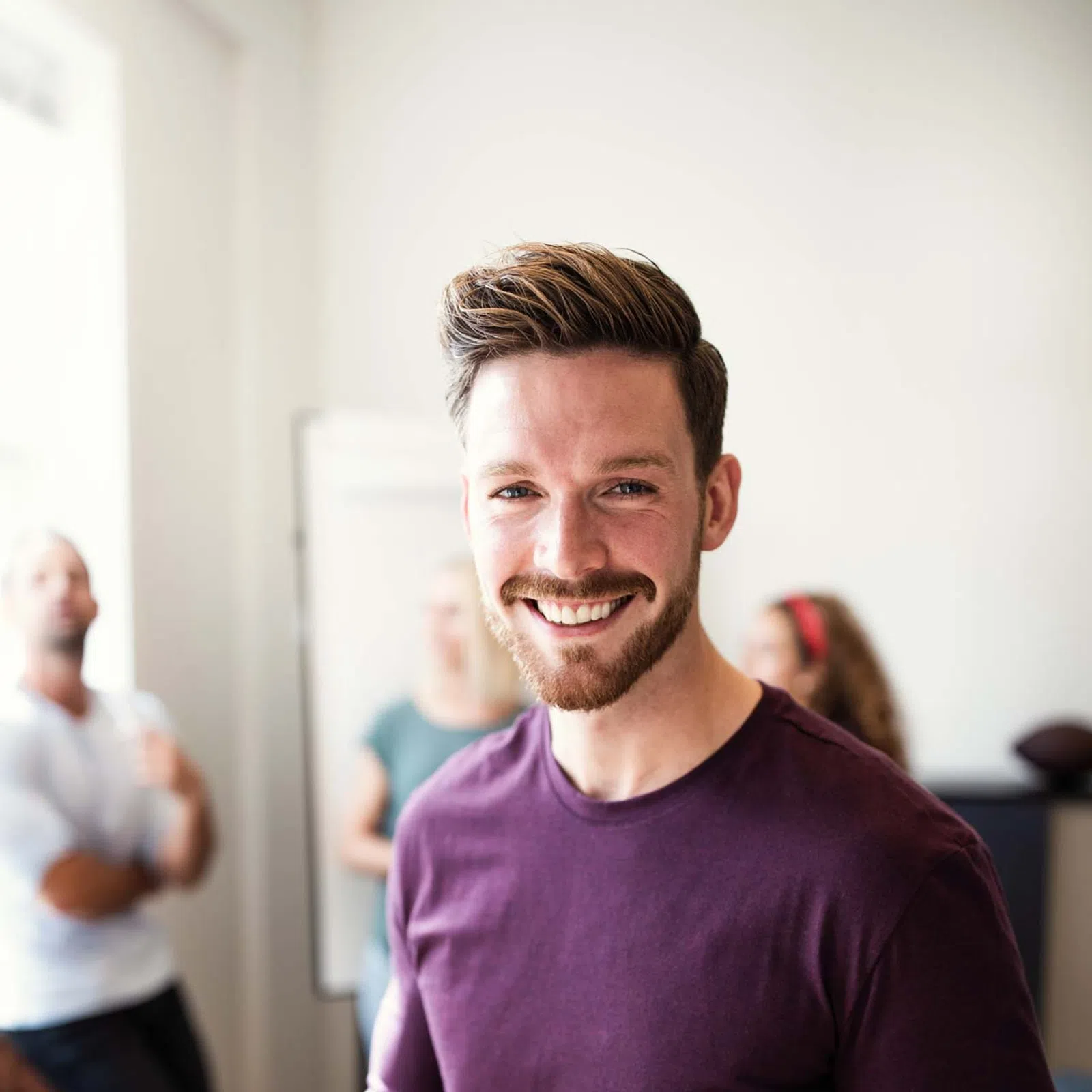 Smiling man in a purple shirt using smartphone with blurred people and office setting in background.