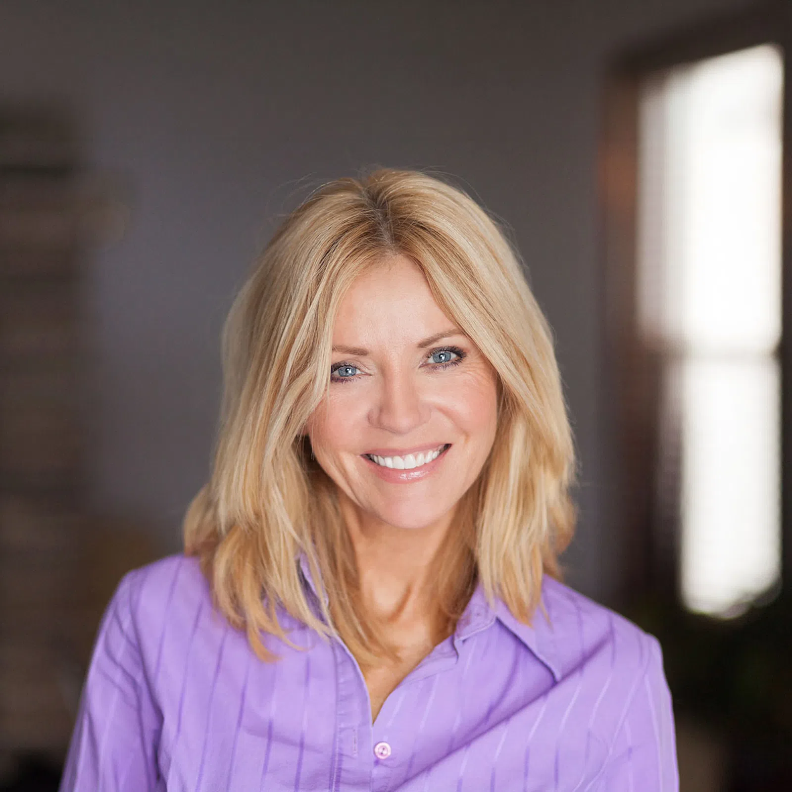 Smiling blonde woman in a purple blouse standing indoors with natural light from window behind her