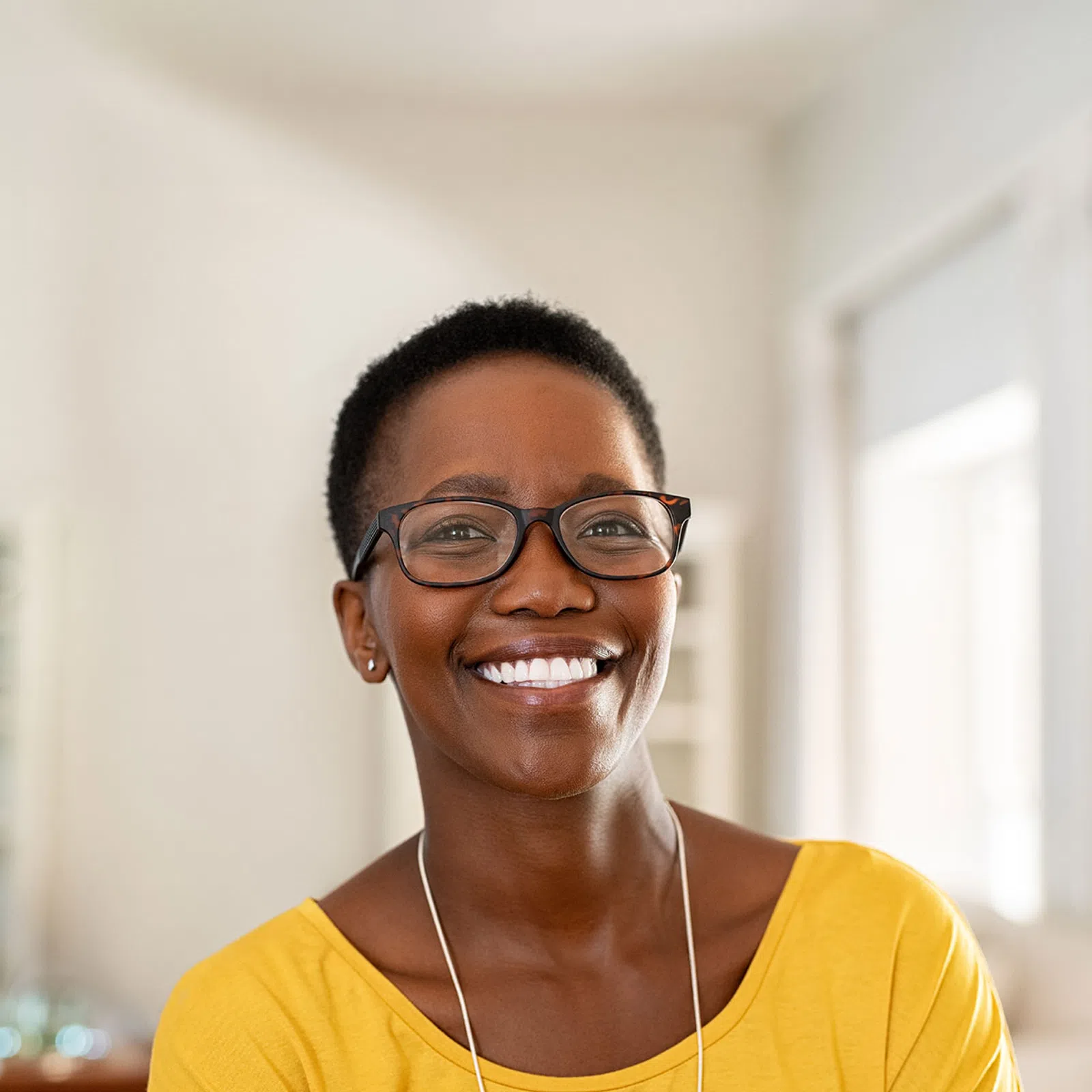 Smiling woman with short hair and glasses wearing a yellow top in a bright, modern room.