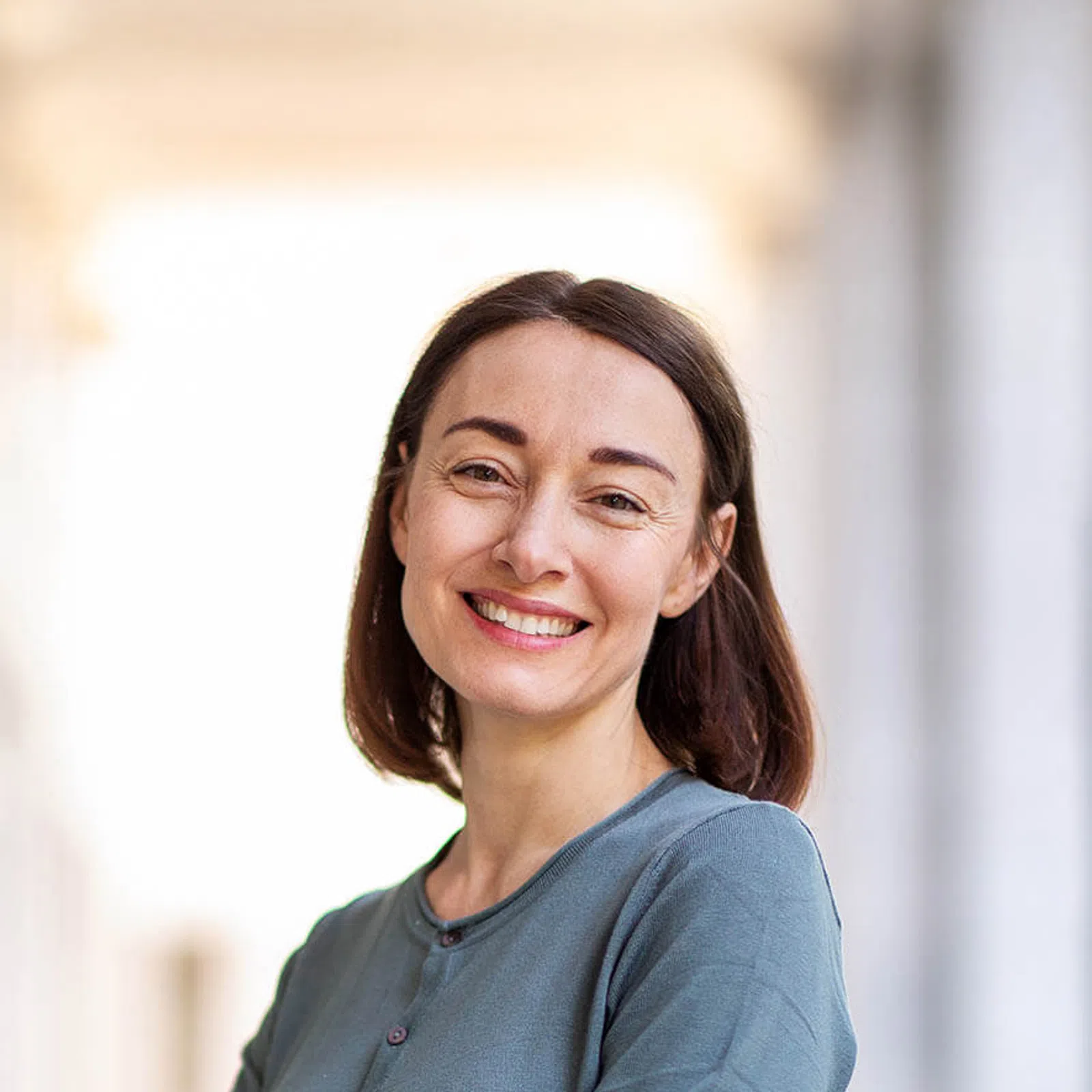 Smiling woman with medium-length brown hair wearing a grey top standing in a softly lit corridor.