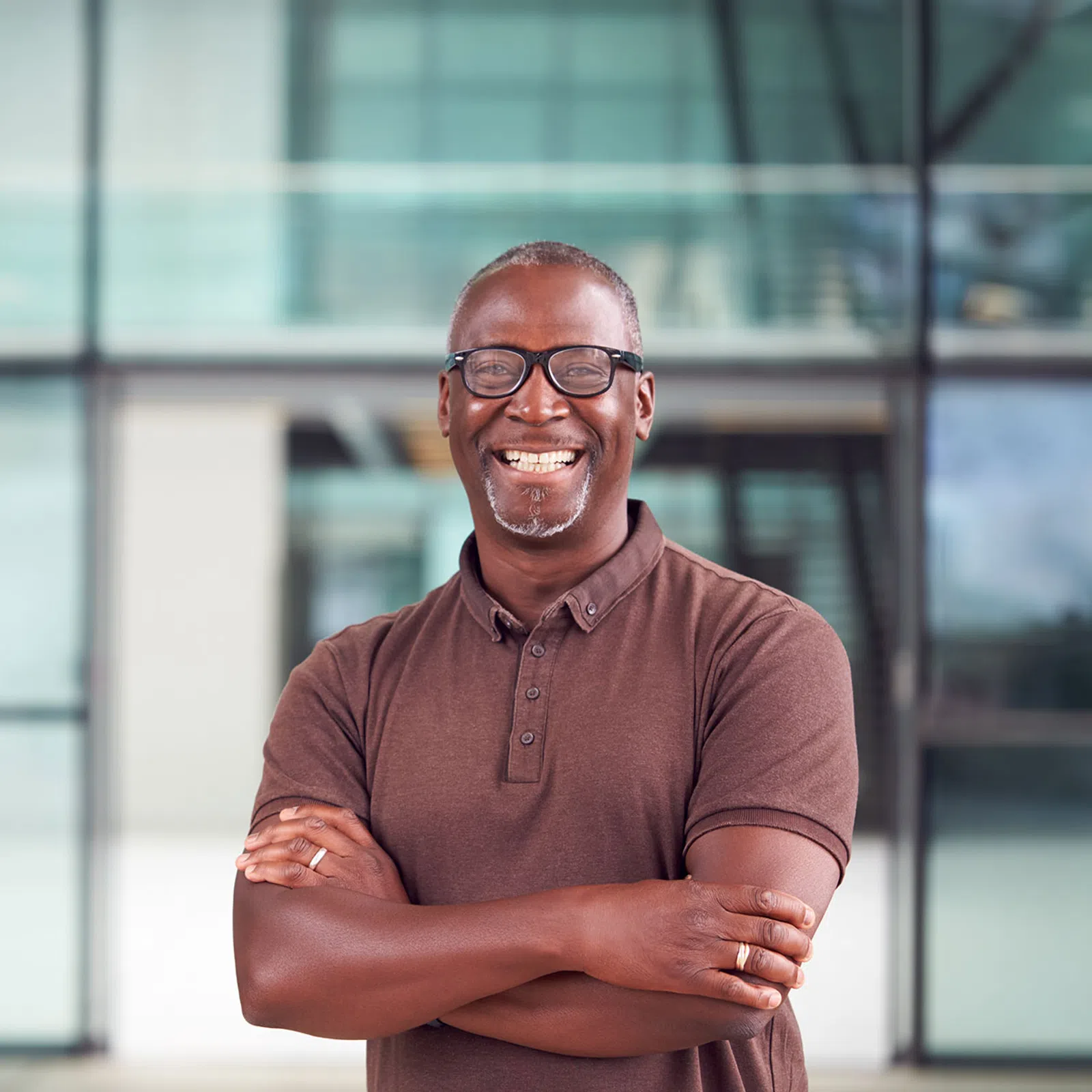 Smiling man wearing glasses and a brown polo shirt standing confidently with arms crossed indoors.
