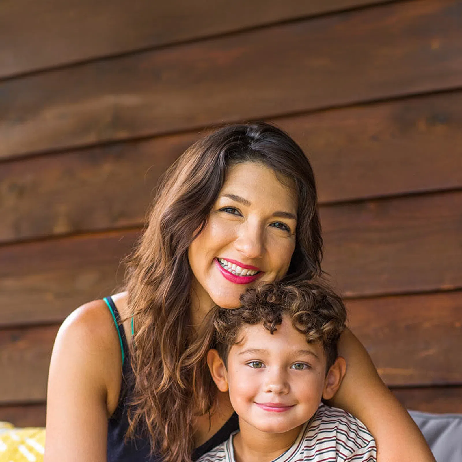 Smiling woman hugging young boy in striped shirt against wooden wall background