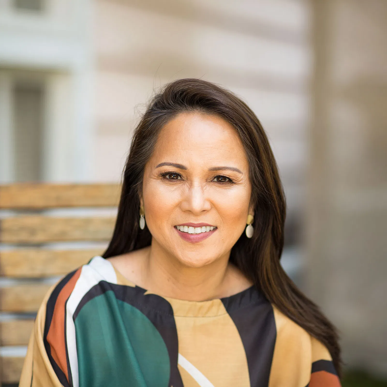 Smiling woman with long dark hair wearing multicolored top sitting on wooden chair outdoors.