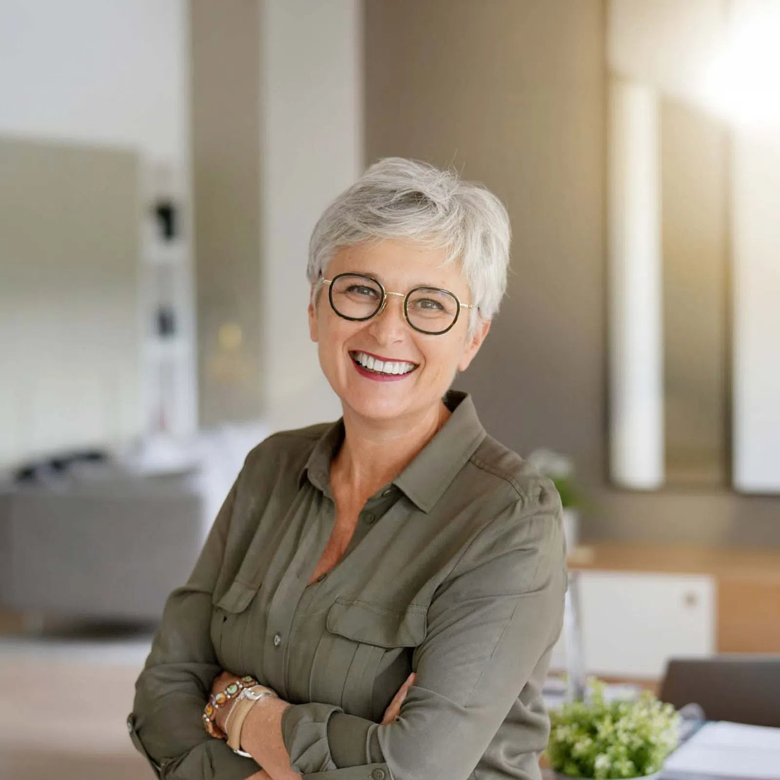 Smiling mature woman with short gray hair and glasses in a casual office setting with crossed arms.
