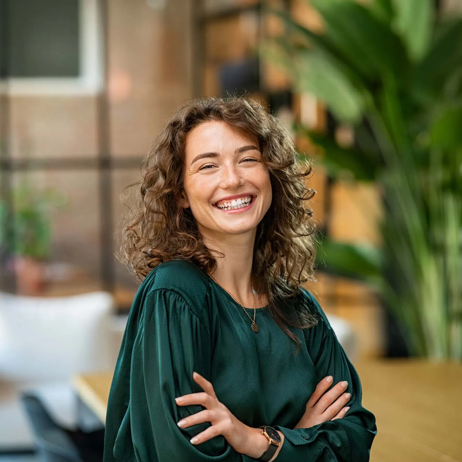 Smiling curly woman in green blouse standing with arms crossed in modern office with plants and blurred background