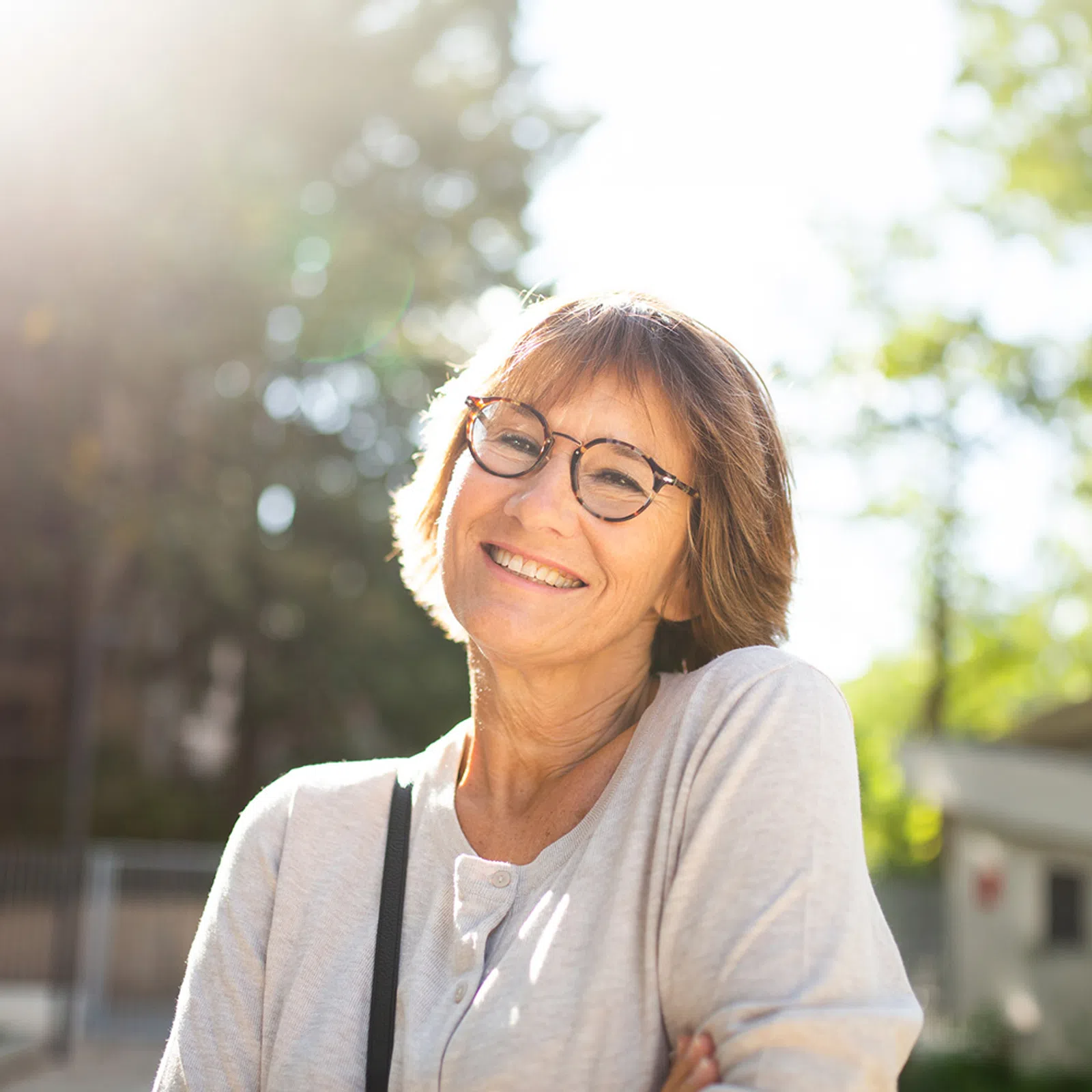 Smiling woman with glasses in casual light sweater standing outdoors in sunlight with trees and buildings in background