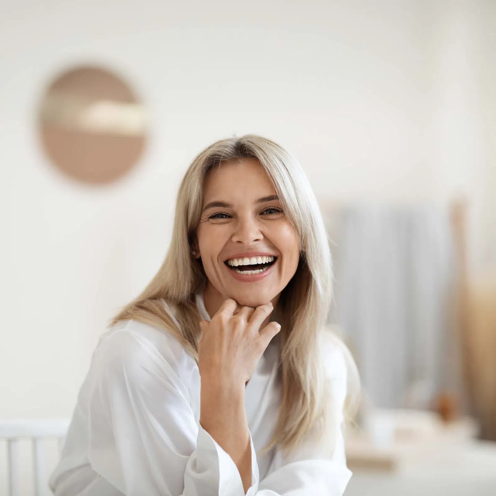 Smiling blonde woman in white blouse sitting indoors with hand on chin against a bright, blurred background.