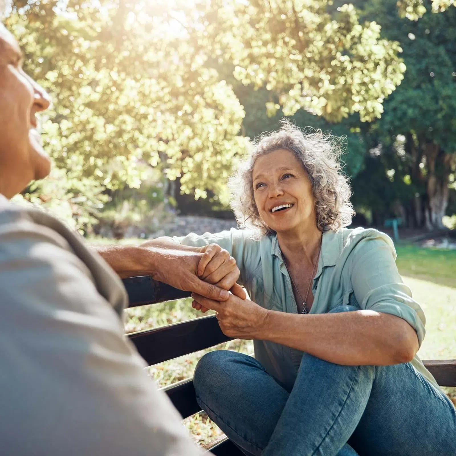 Smiling senior woman holding hands with elderly man on park bench in sunlit green space.