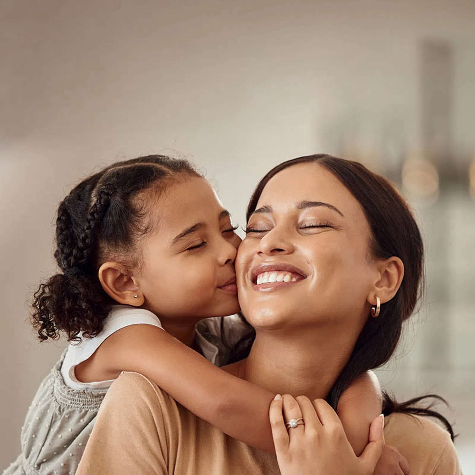 Young girl kisses her smiling mother on the cheek while hugging her tightly in a warm family moment.
