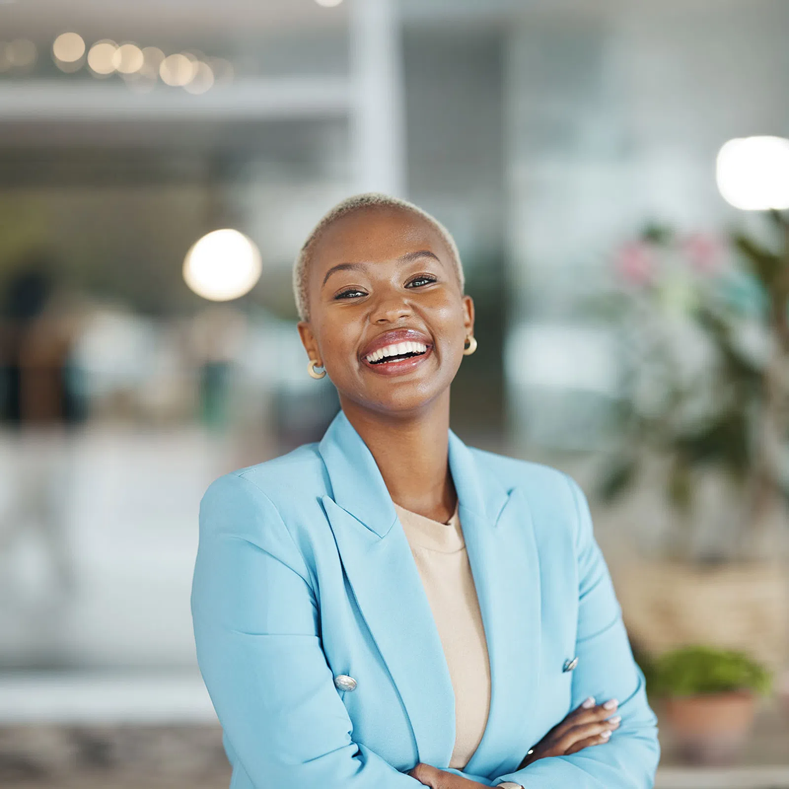 Smiling confident woman in blue blazer standing with arms crossed in modern office space.