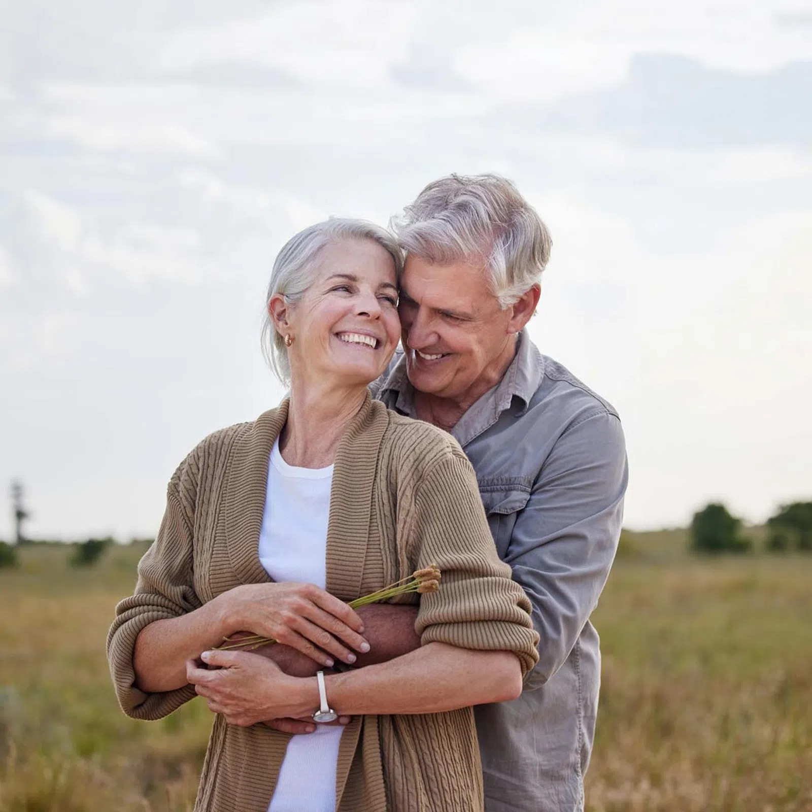 Happy senior couple embracing outdoors in a scenic field during daytime with smiles and casual clothes.