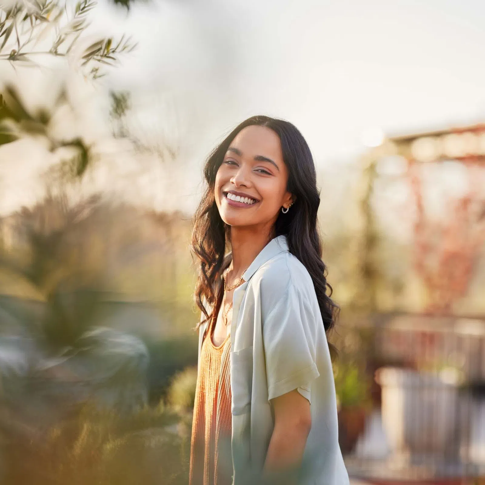 Smiling young woman with long dark hair standing outdoors in soft sunlight near greenery and blurred background.