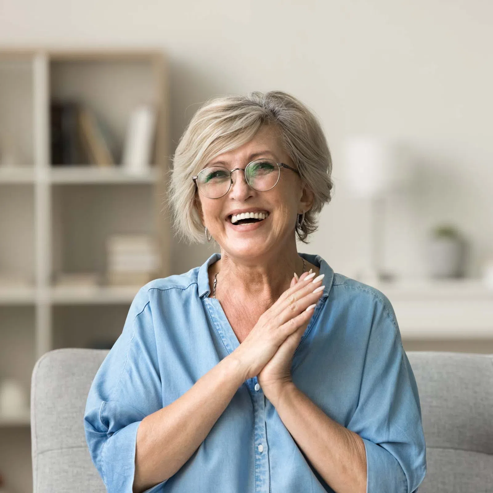 Smiling elderly woman with glasses sitting on a gray sofa in a bright living room with bookshelves.