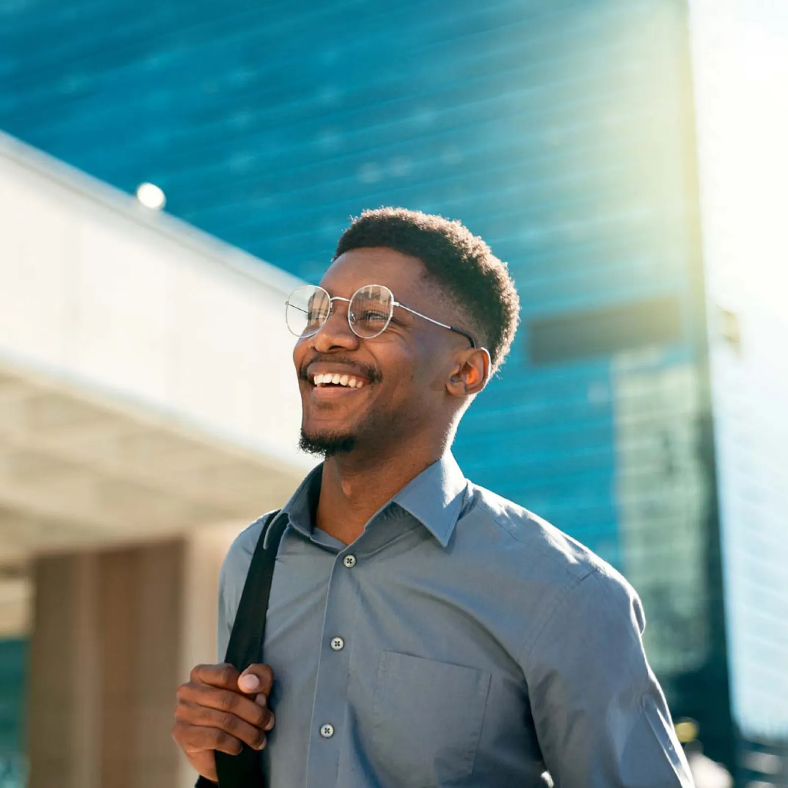 Smiling young man wearing glasses and carrying a backpack walks outdoors in urban setting on sunny day.