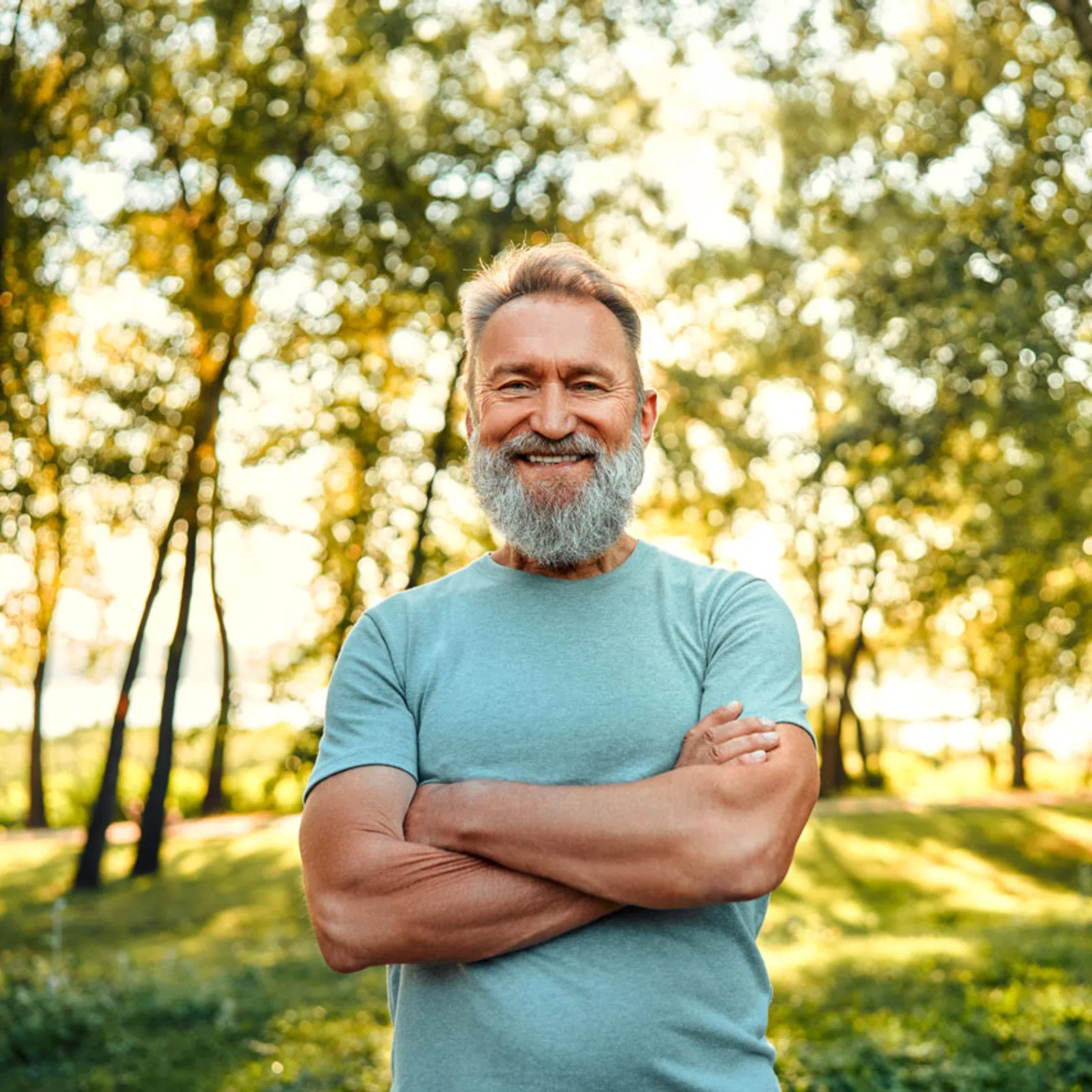 Smiling middle-aged man with gray beard and crossed arms standing outdoors in sunlit park with trees.
