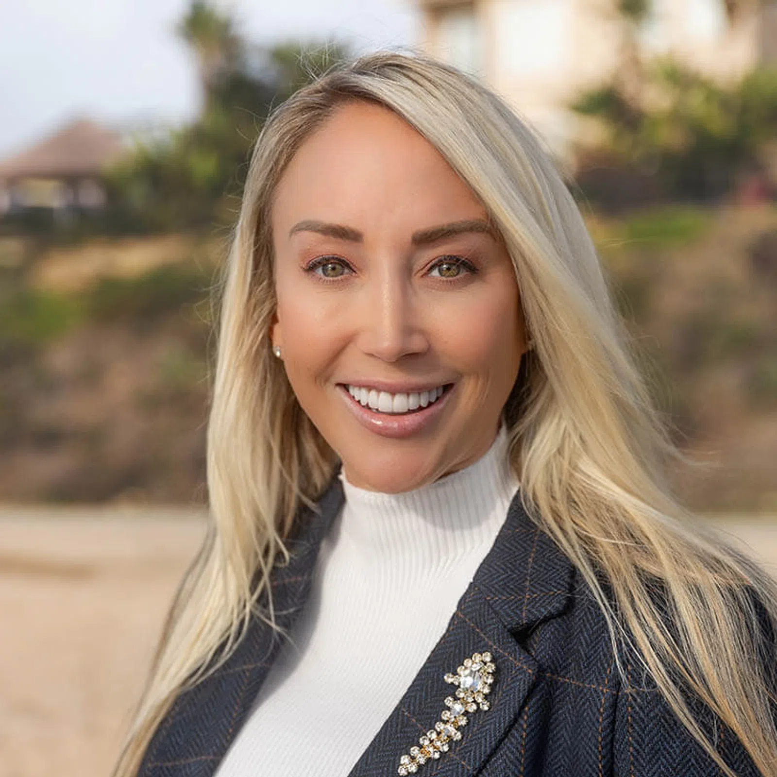 Smiling blonde woman in white turtleneck and dark blazer with decorative brooch outdoors in natural setting.