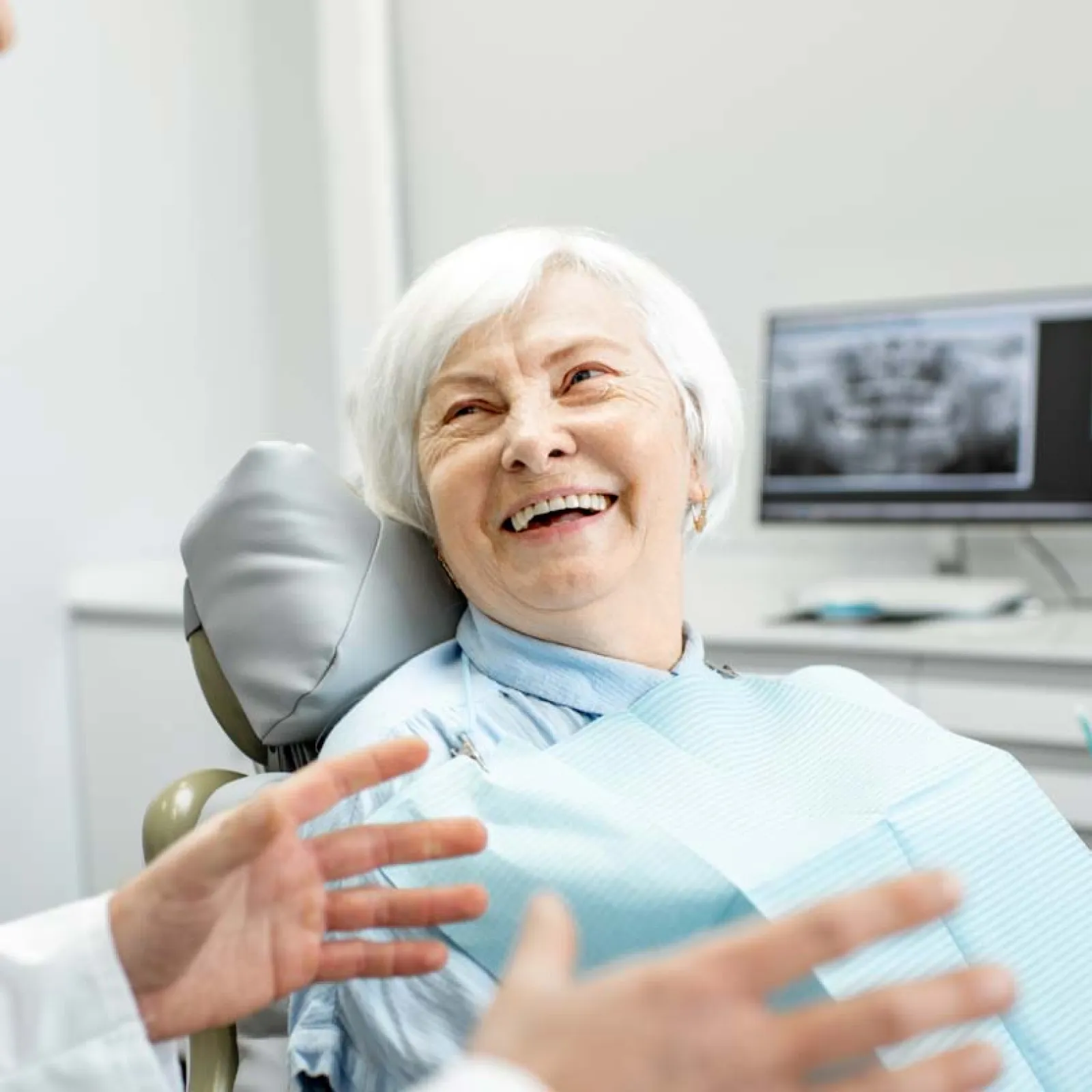 Elderly woman smiling in dental chair during consultation with dentist and dental x-ray on screen behind.