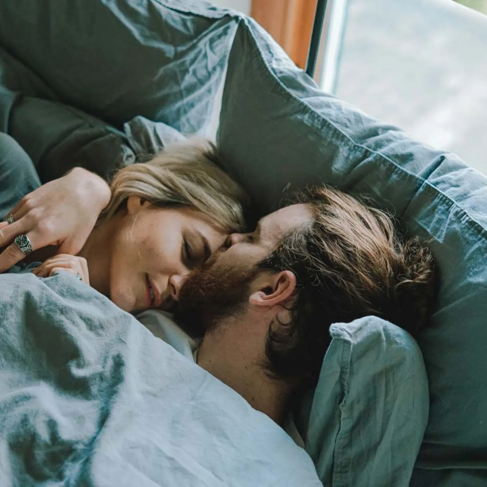 Couple cuddling closely in bed under blue-gray sheets beside a window, sharing an intimate moment.