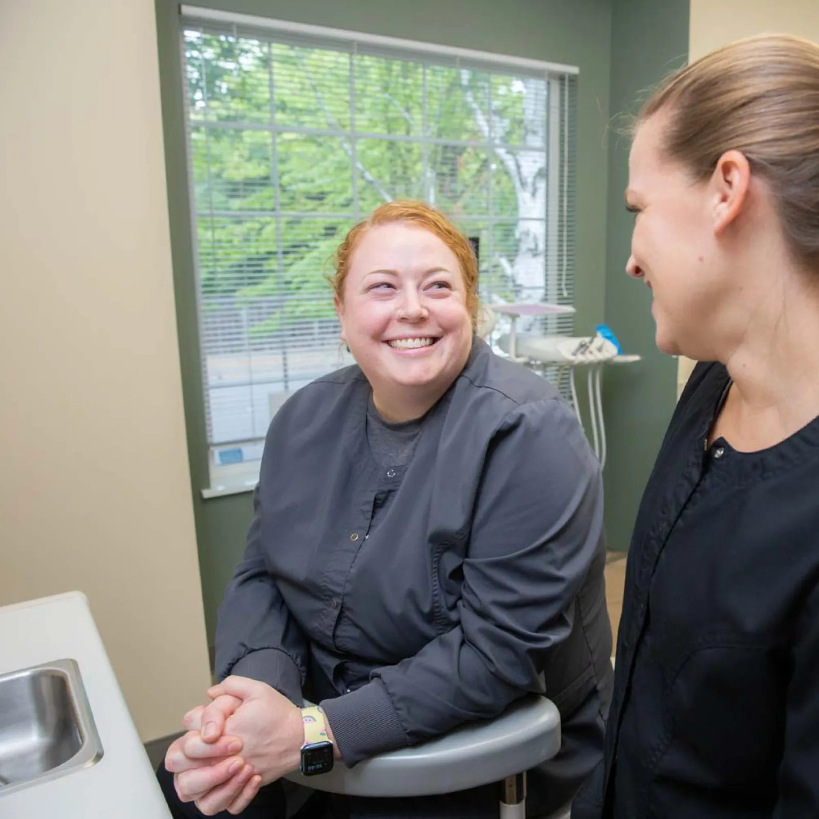 Two female healthcare professionals smiling and conversing in a bright medical office with window and equipment.