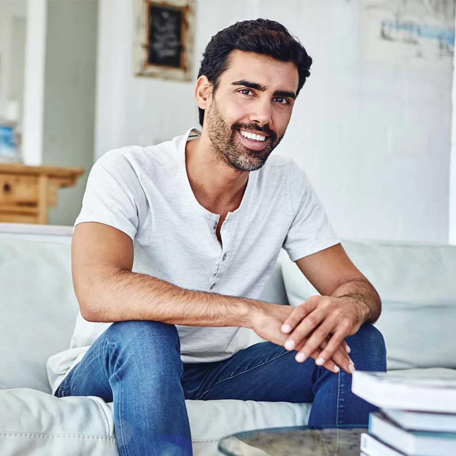 Smiling man with beard sitting on couch wearing white shirt and blue jeans in bright living room