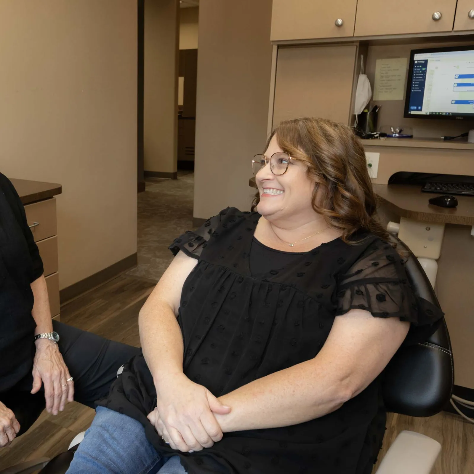 Two women smiling and chatting in a modern office with beige walls and a computer workstation.