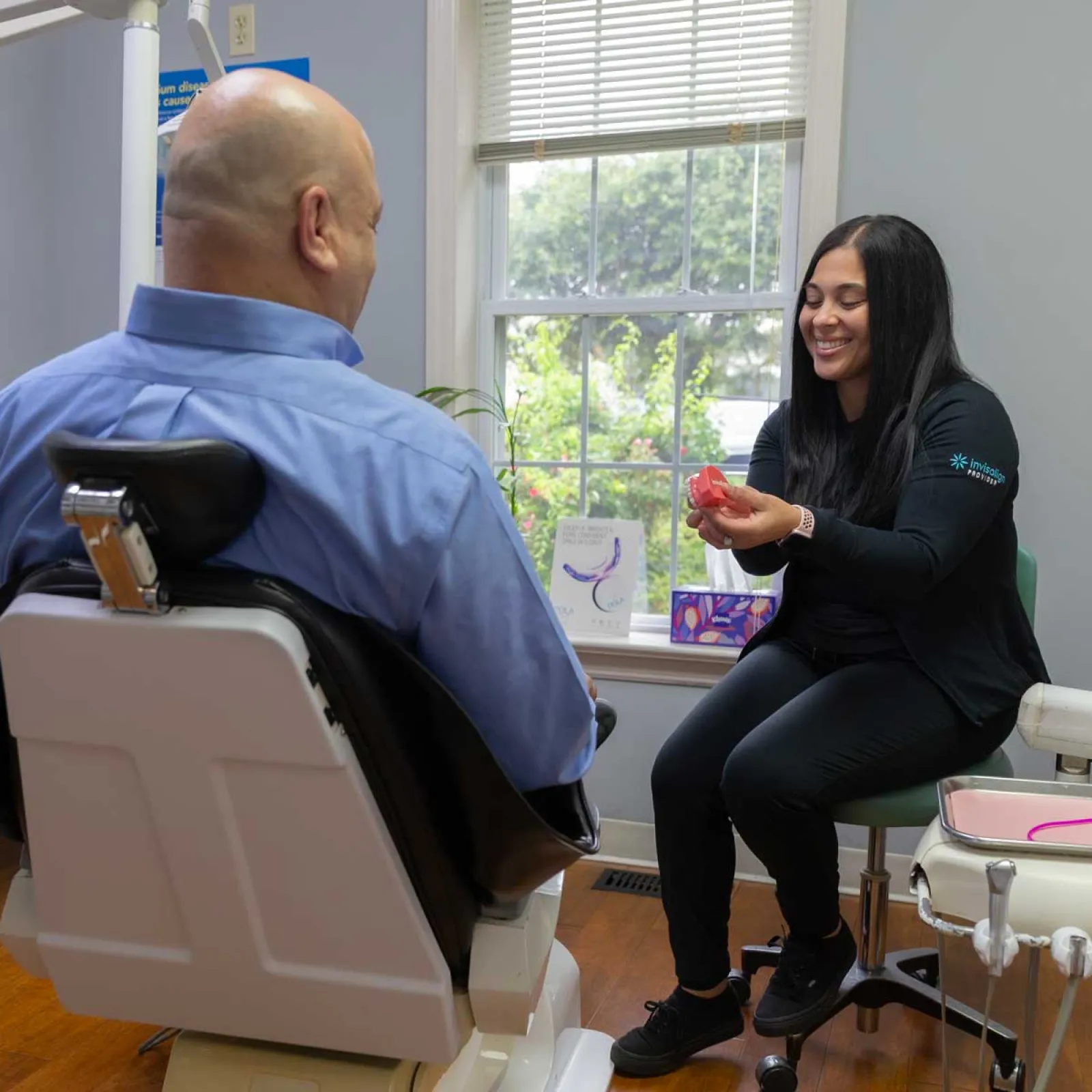 Dental professional explaining teeth aligner to male patient in modern clinic with large window lighting.