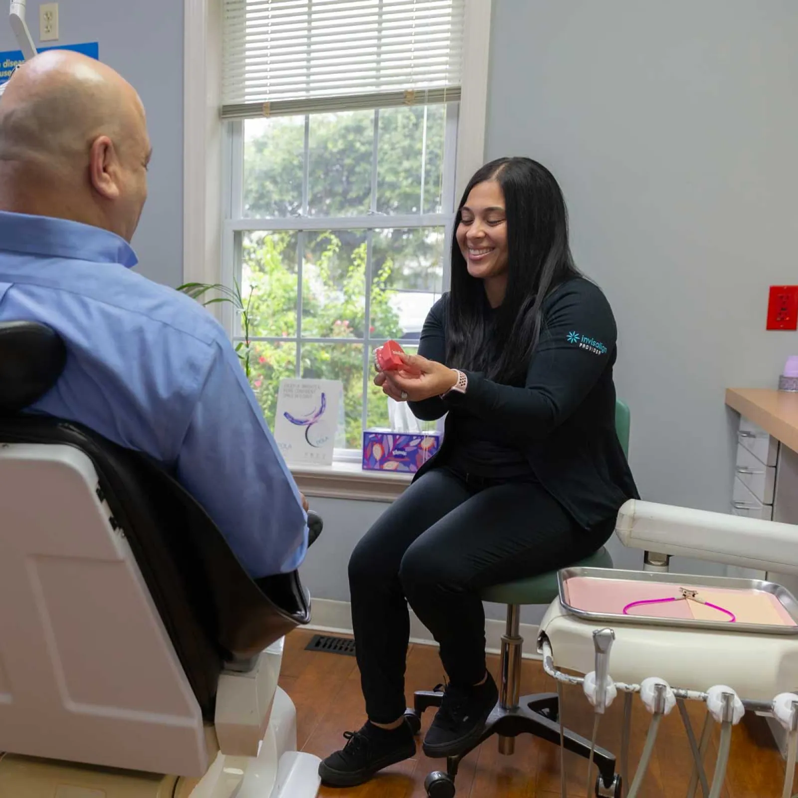 Dental professional explaining teeth aligner to male patient in modern clinic with large window lighting.