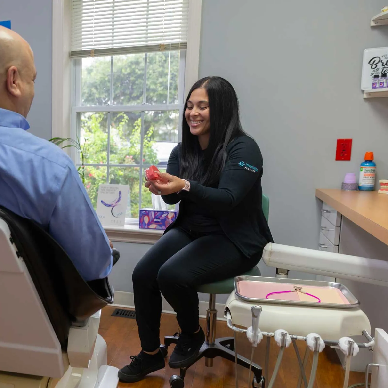 Dental professional explaining teeth aligner to male patient in modern clinic with large window lighting.