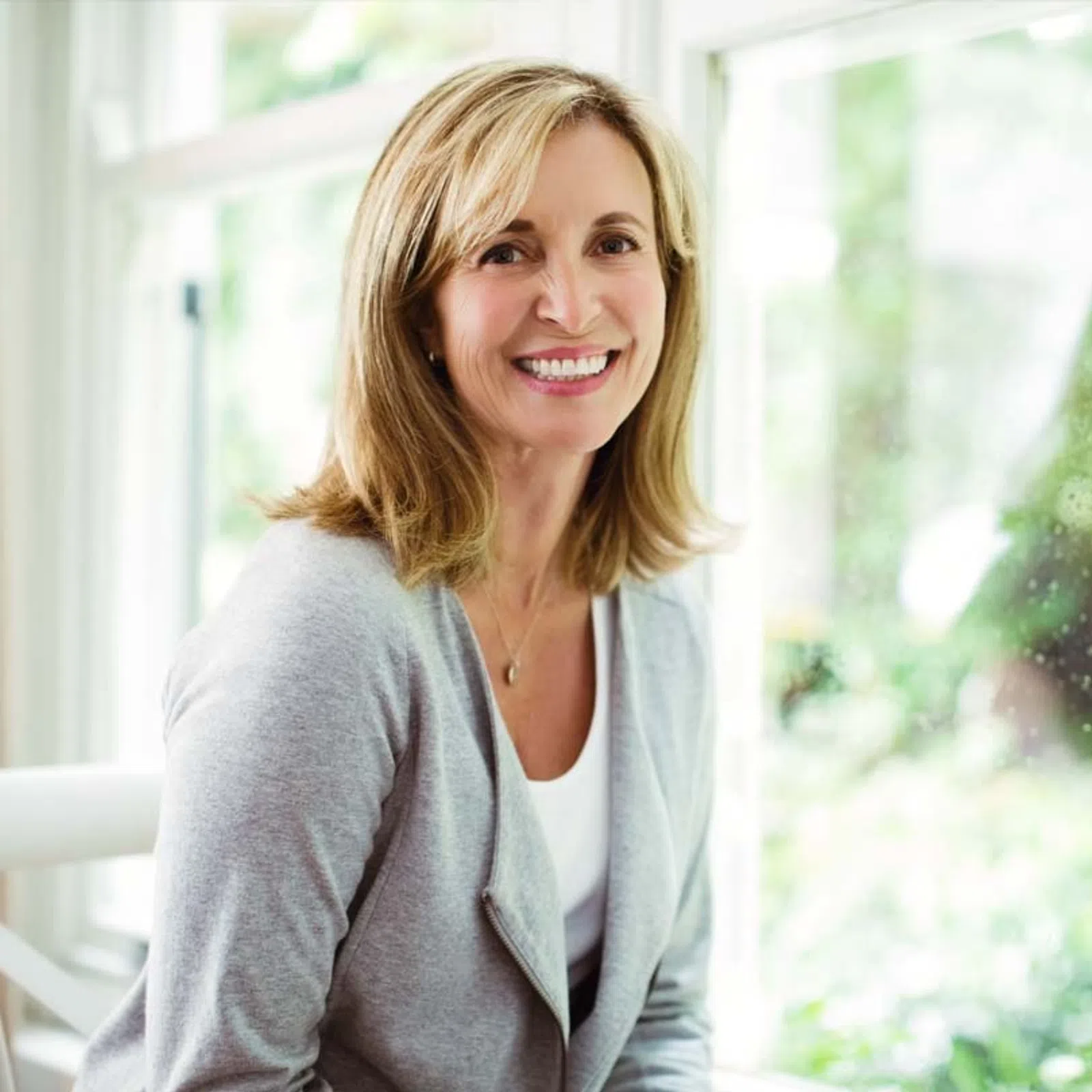 Smiling middle-aged woman with blonde hair wearing a gray cardigan sitting by a bright window indoors.