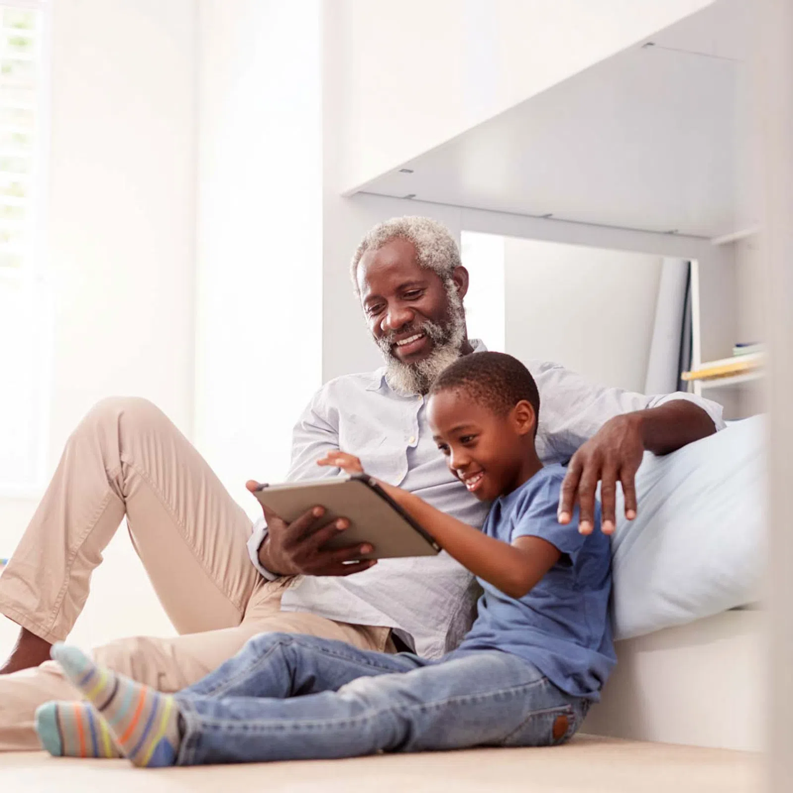 Grandfather and grandson sitting on floor using a tablet together in a bright living room.