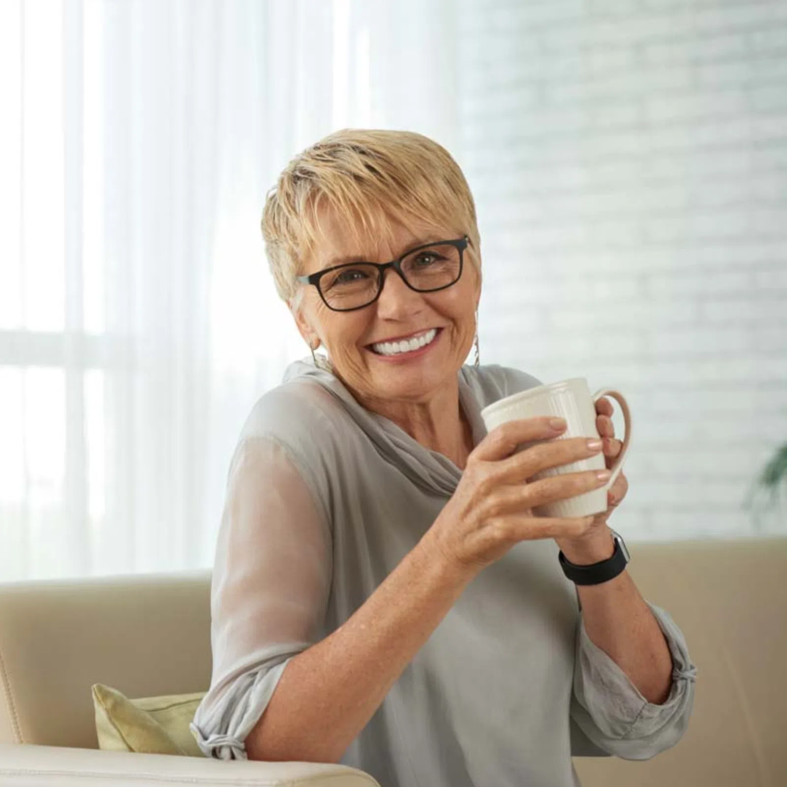 Smiling mature woman with glasses holding a coffee mug while sitting on a couch in a bright room