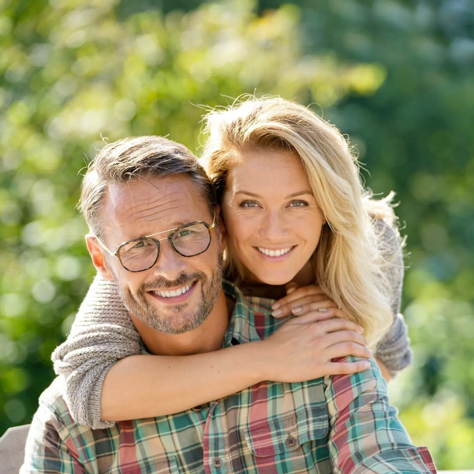 Happy middle-aged couple outdoors, woman hugging man from behind with green blurred background.