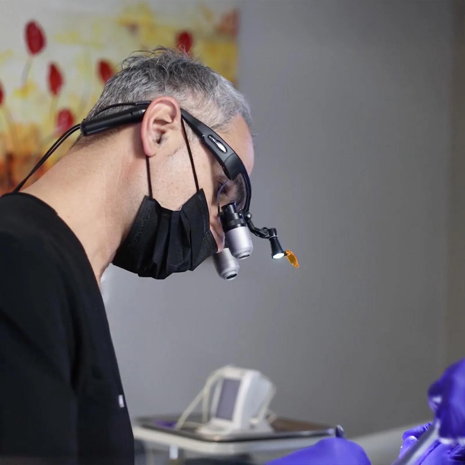 Medical professional wearing magnifying glasses and black mask performing a precise procedure in a clinical setting.