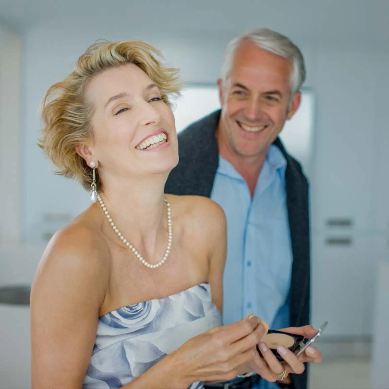 Happy mature woman applying makeup with smiling man in background in bright modern room.