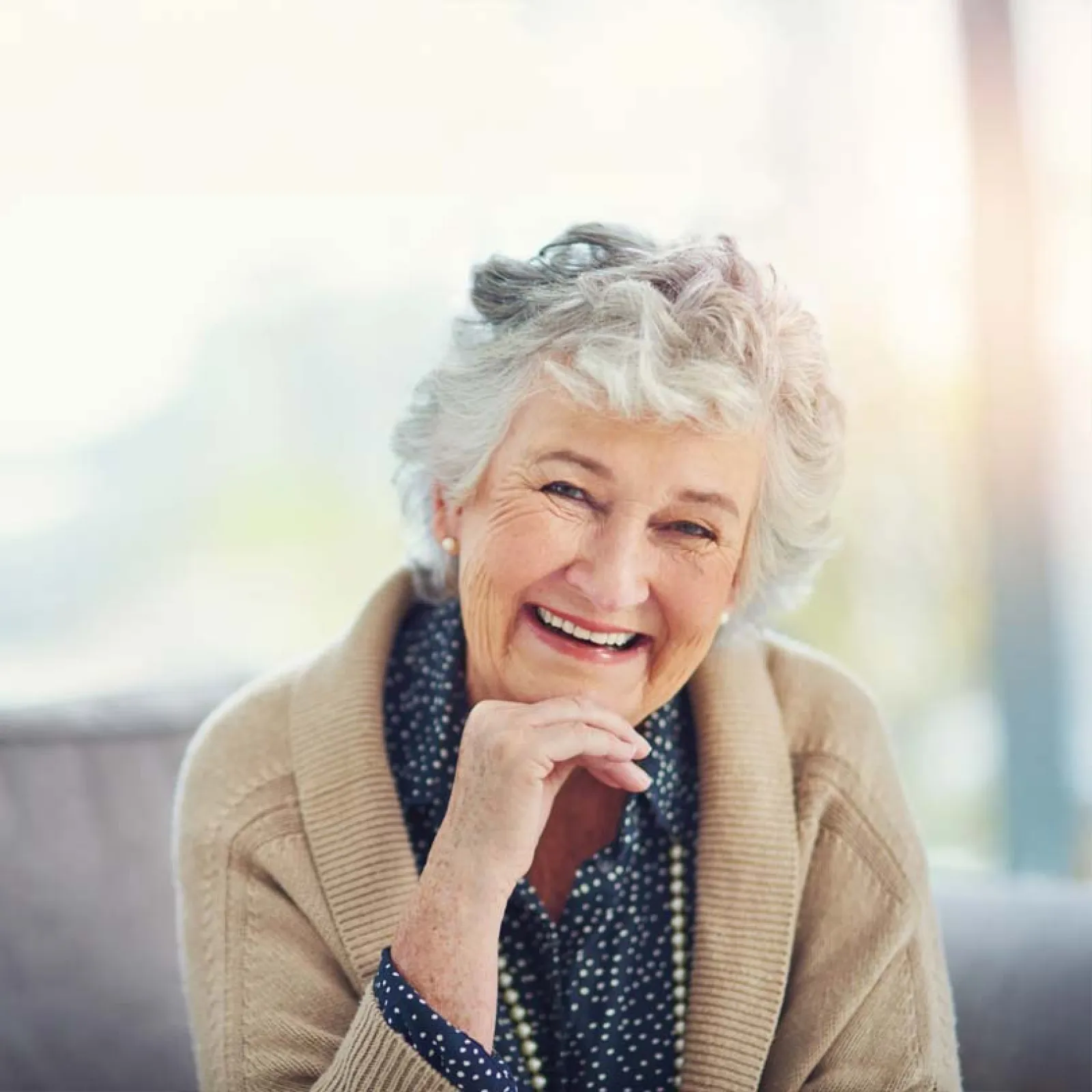 Smiling elderly woman with curly gray hair wearing a beige cardigan and patterned blouse indoors.