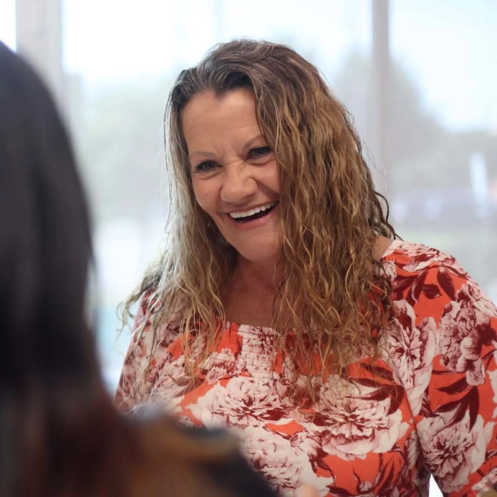 Smiling woman with curly hair wearing a red and white floral top engaged in conversation indoors