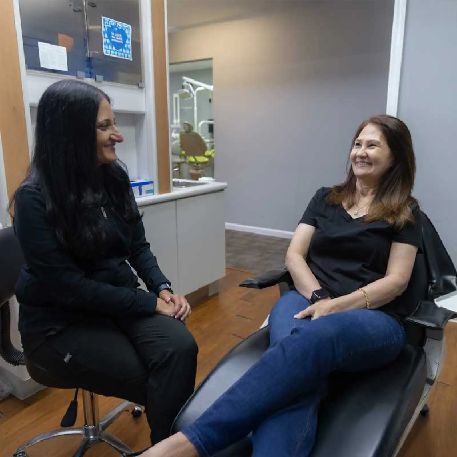 Two women smiling and talking in a dental clinic, one seated in a dentist chair and the other on a stool.