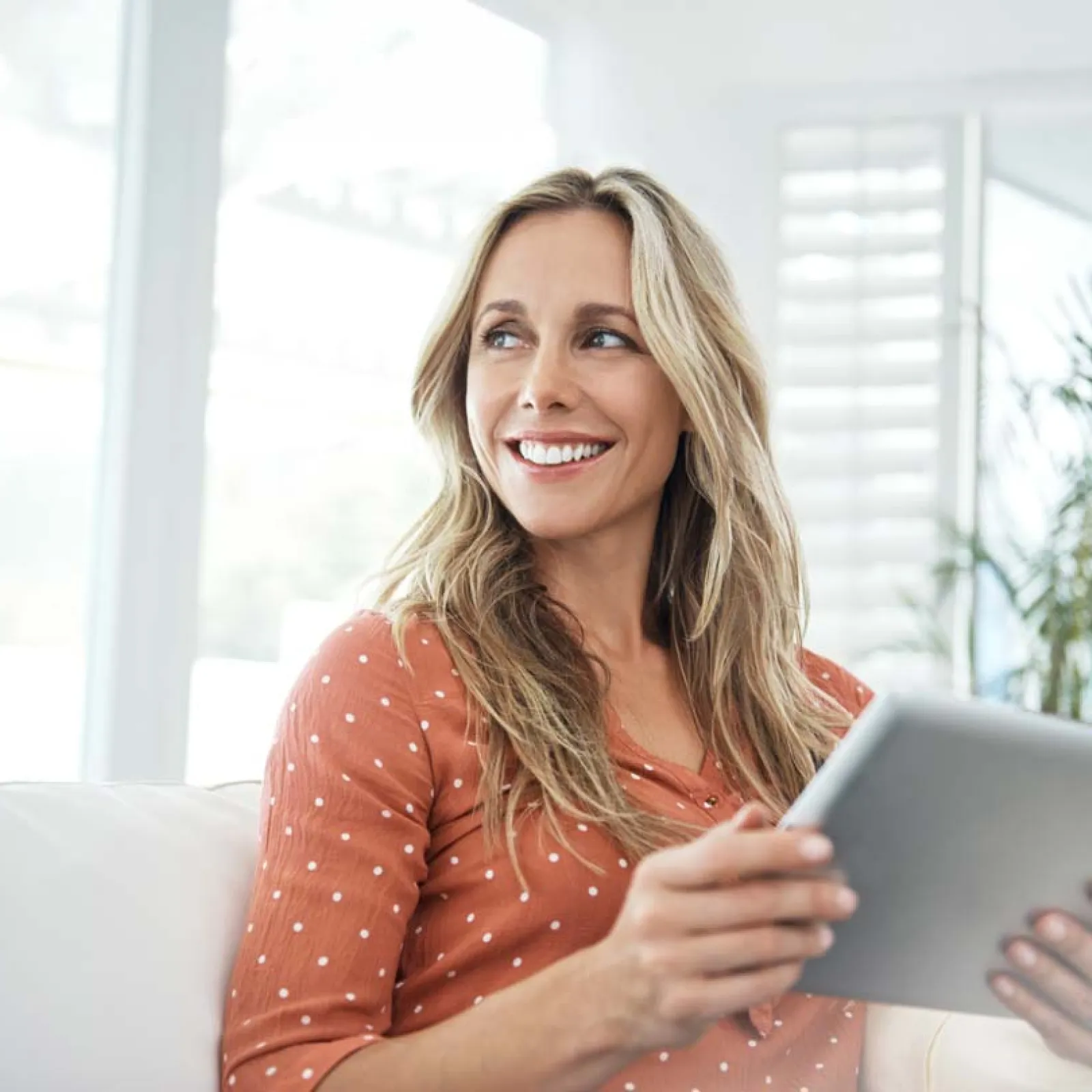 Smiling woman with blonde hair holding a tablet while sitting on a white couch in a bright room.