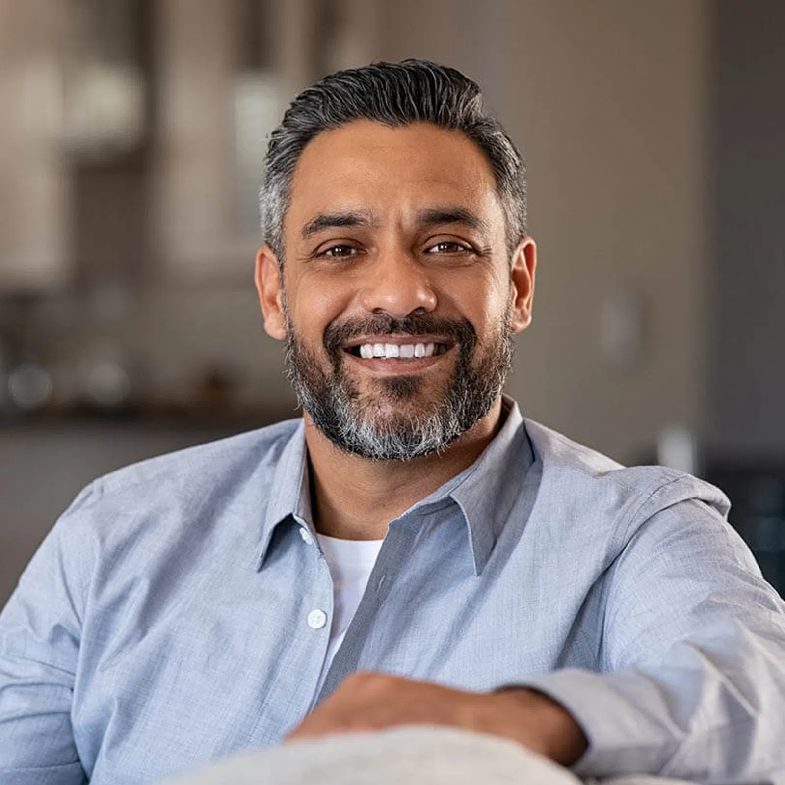 Smiling middle-aged man with salt-and-pepper beard wearing a light blue shirt indoors, looking at camera.