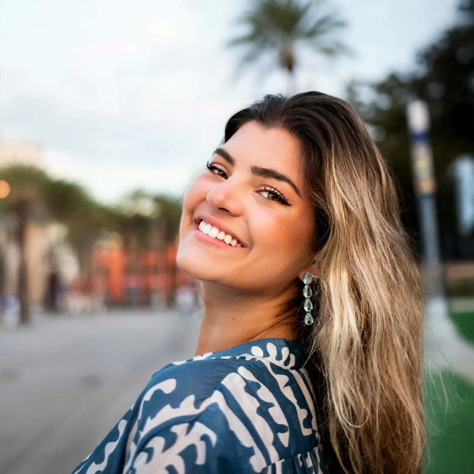 Smiling young woman with long hair and blue patterned shirt outdoors with blurred background.