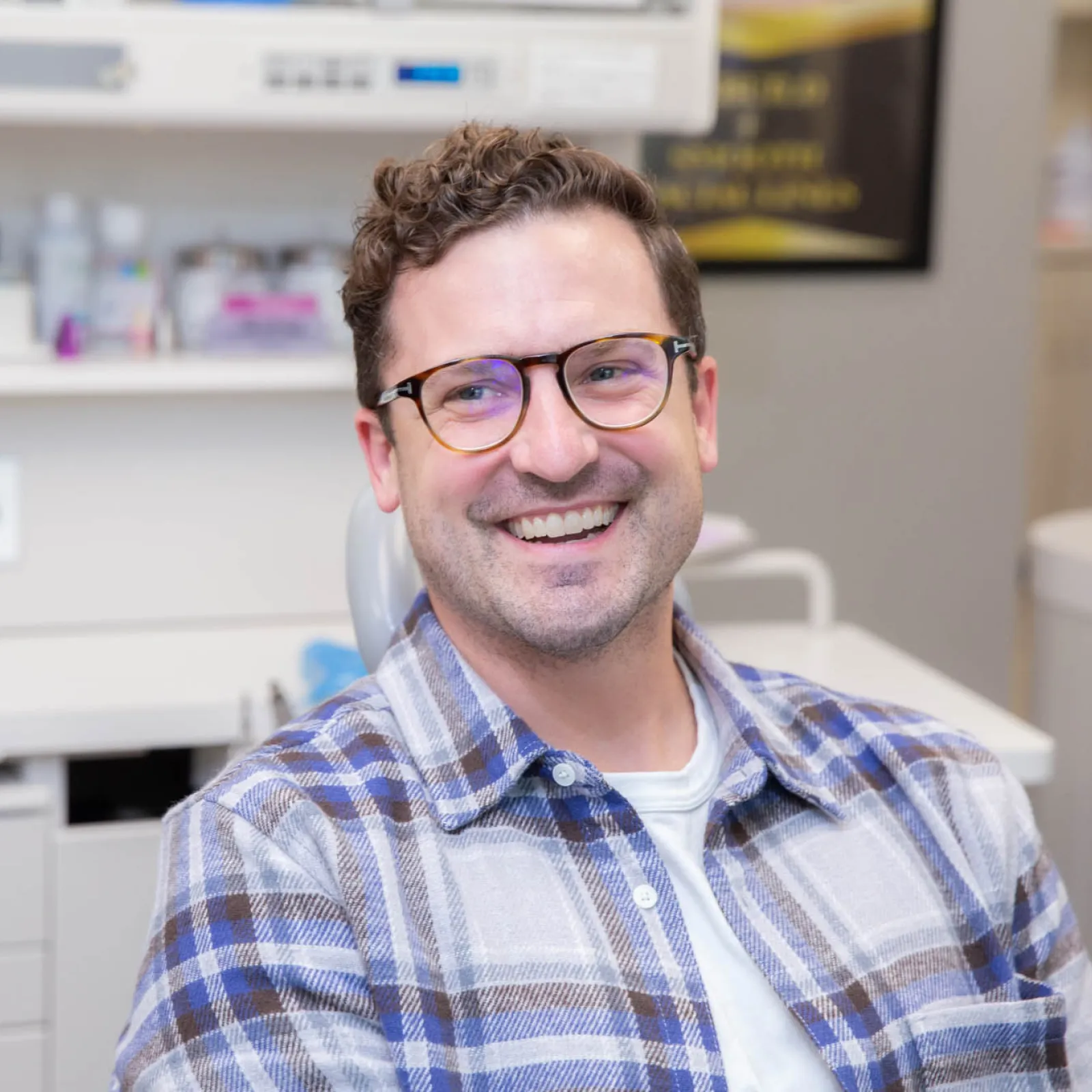 Smiling man wearing glasses and plaid shirt sitting in a modern dental office talking to a dentist.