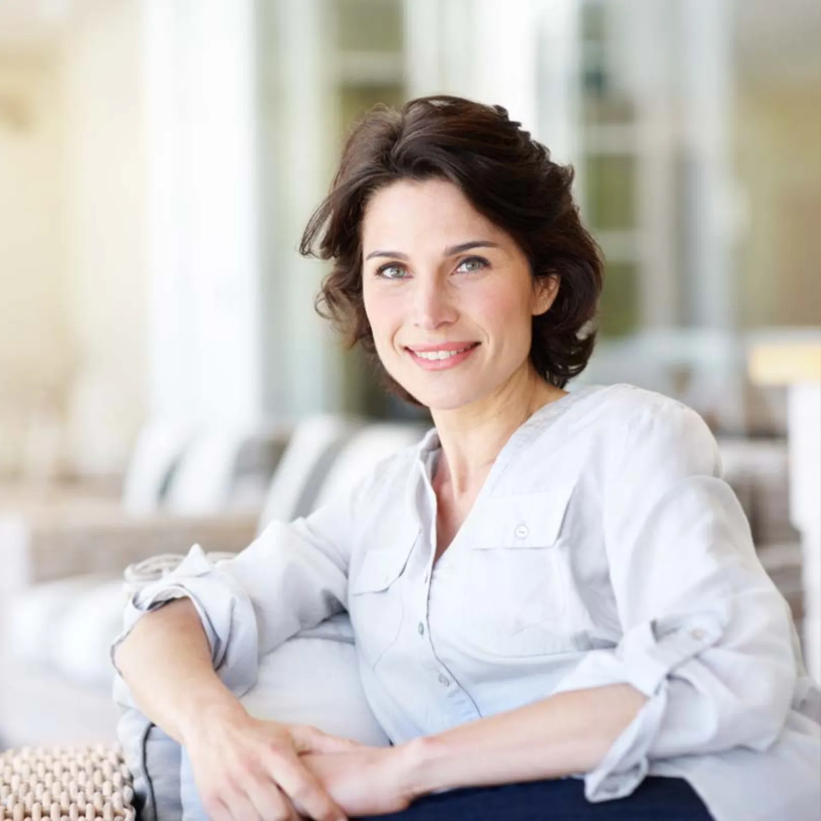Smiling woman with short brown hair in white blouse sitting on a couch in a bright, modern room.