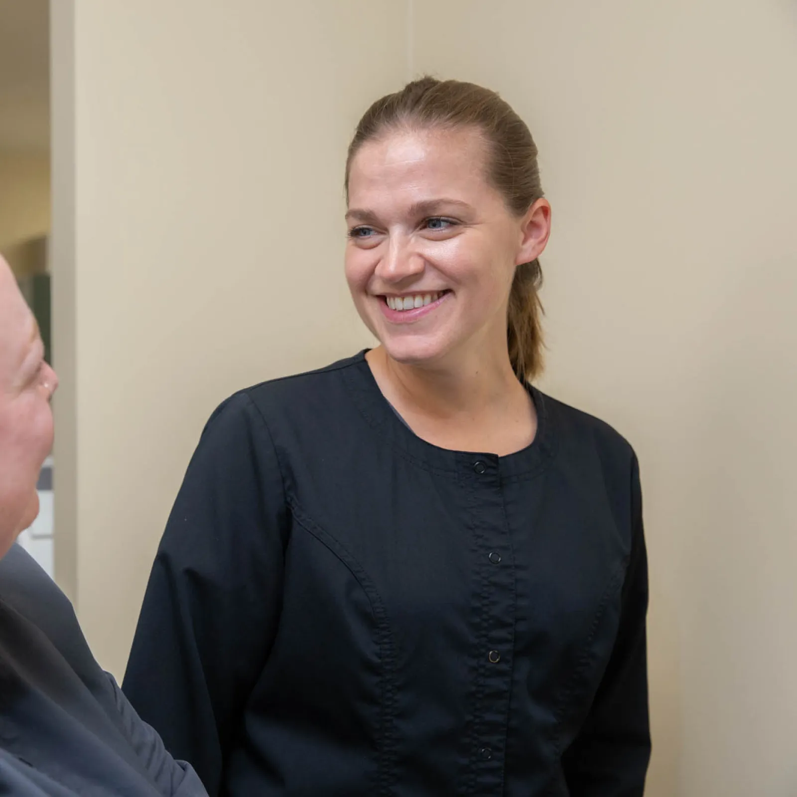 Two women in black uniforms smiling and talking in a neutral indoor setting with beige walls.