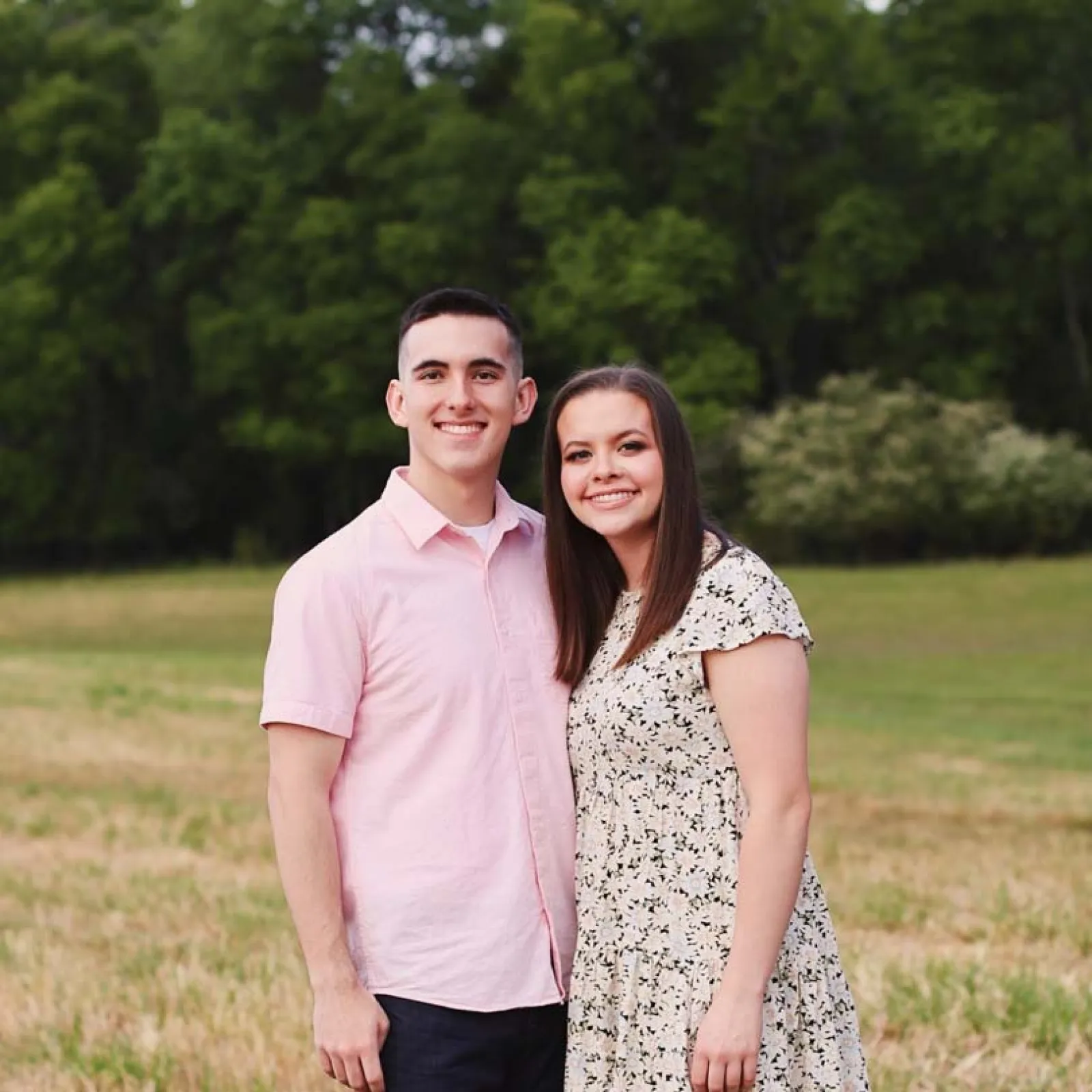 Smiling young couple standing close outdoors in a grassy field with trees in the background