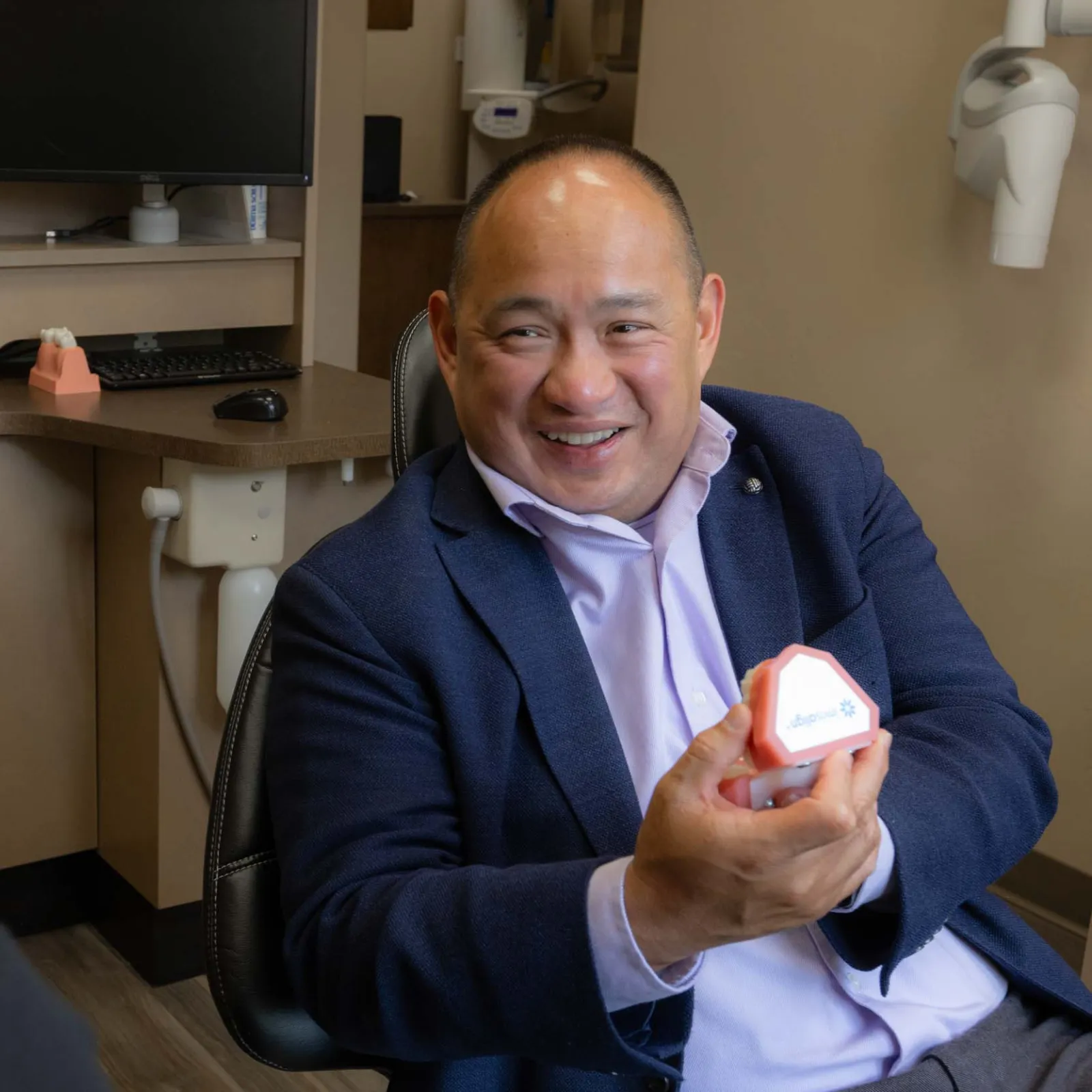 Dentist in office showing dental mold to patient during consultation with chair and equipment in background